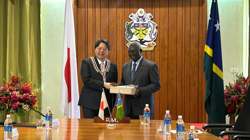 Japanese foreign minister and Solomon Islands PM hold white box and smile at end of table.