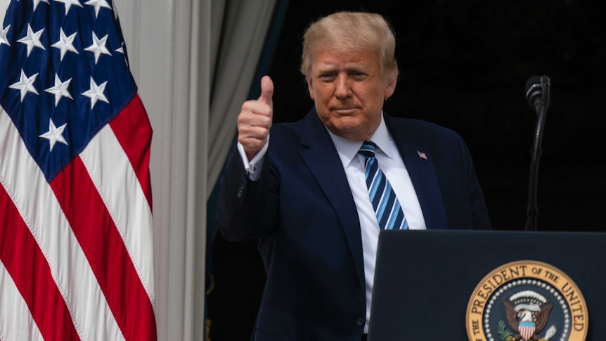 A man in a navy suit gives a thumbs up as he stands behind a lectern near a US flag