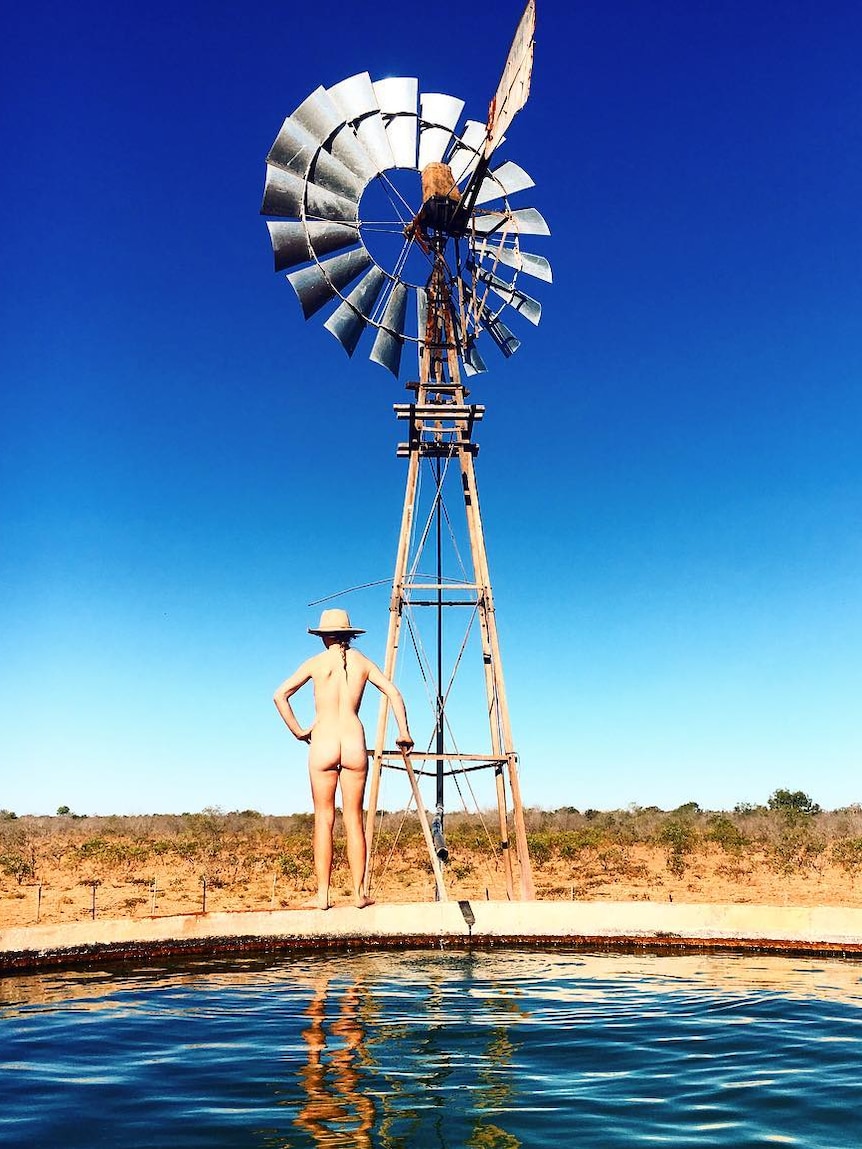 A naked woman stands on the edge of a tank near a windmill