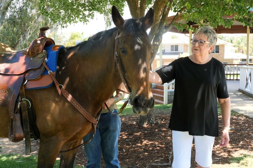 A woman with glasses pats a horse's nose at a park.