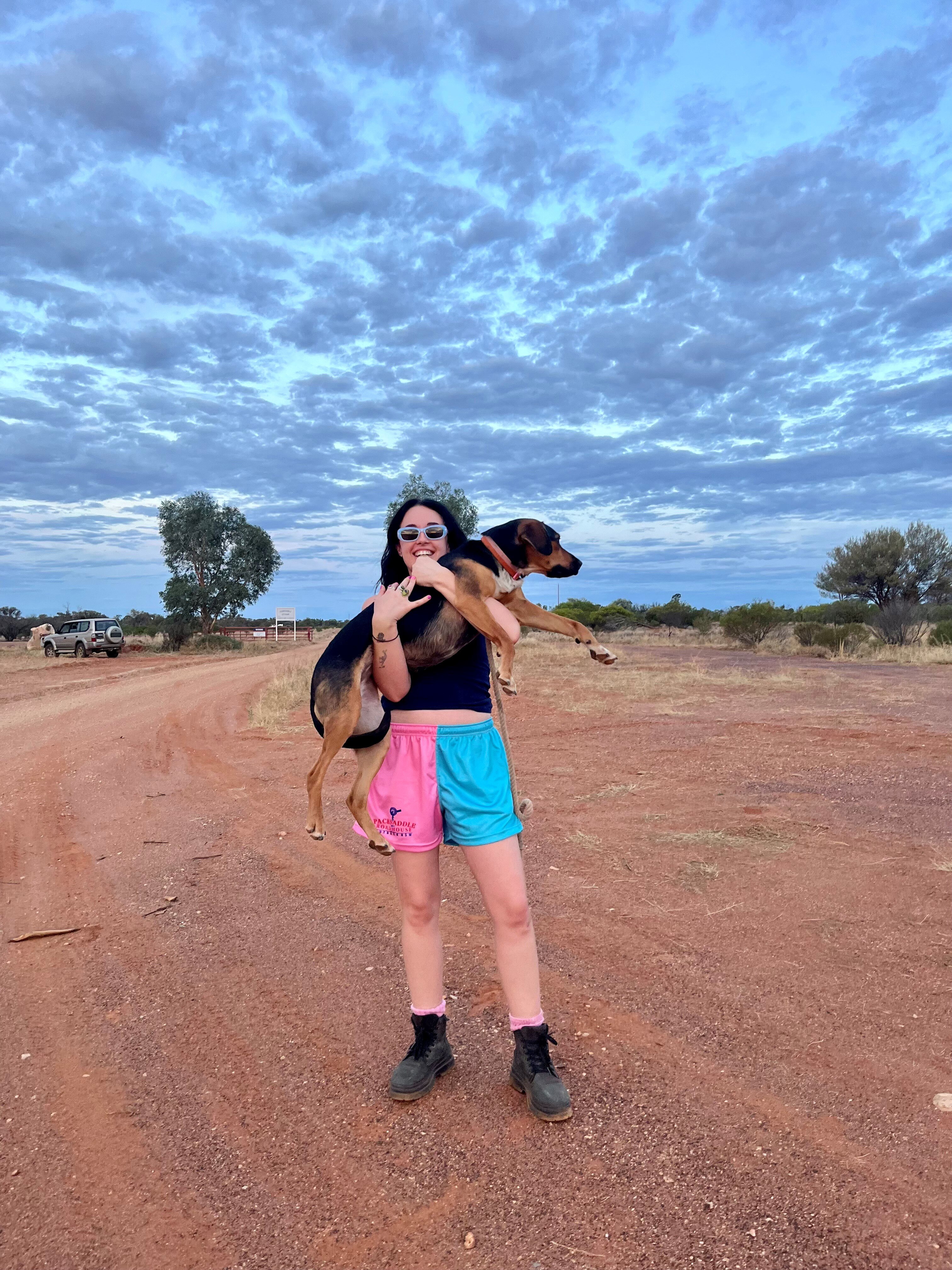 Una mujer con pantalones cortos multicolores sostiene un kelpie en sus brazos: tierra roja, suelo desnudo, árboles a lo lejos, nubes azules.