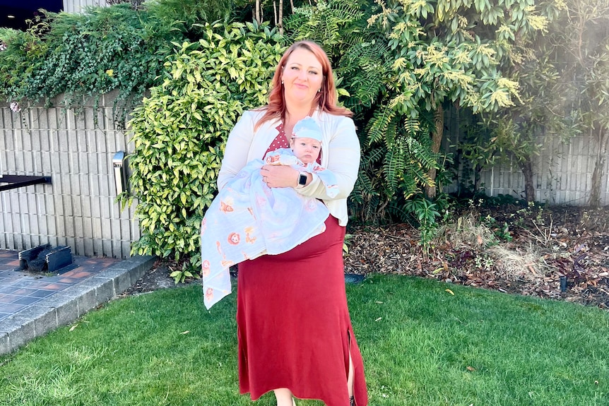 A woman in a red dress is standing outside a building holding a new baby.