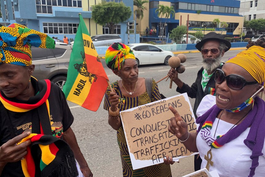 A group of protesters dressed in traditional Jamaican colours gather on a street. One is holding a sign that says #60reasons