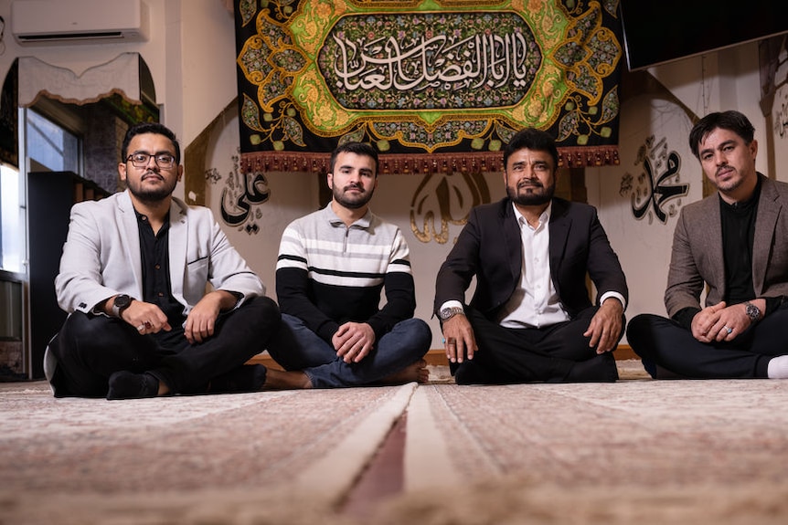 Four men sit on a carpet with Arabic script behind them.
