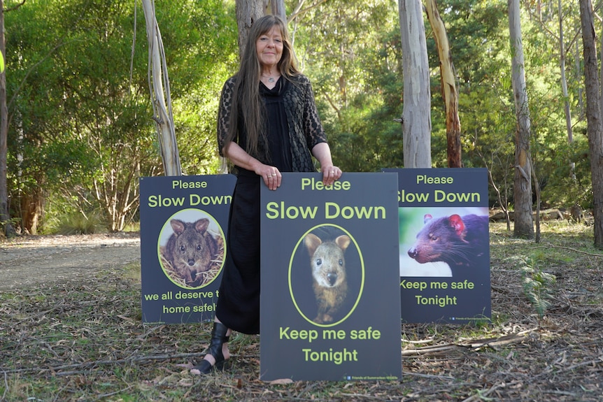 Ruth is standing in eucalyptus forest holding signs she made featuring native animals urging drivers to slow down.