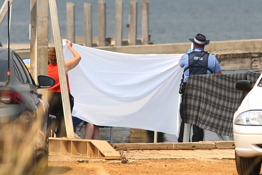Police hold up a white sheet next to a beach, with parked cars in the foreground.
