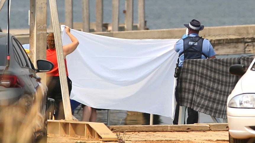 Police hold up a white sheet next to a beach, with parked cars in the foreground.