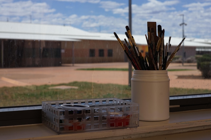 Paint brushes in a ceramic jar in a window. A plastic box with holes has colour pots, low orange buildings in the distance.