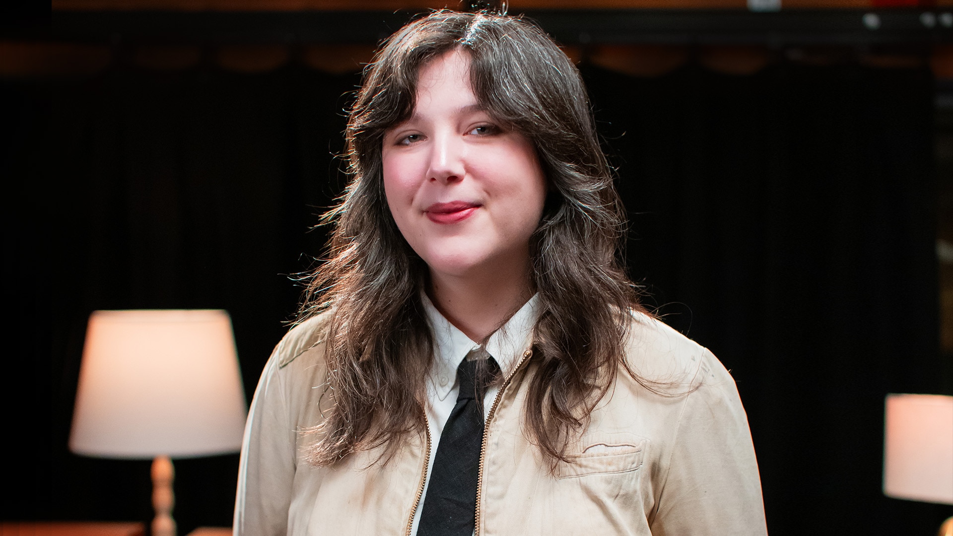 Lucy Dacus stands and smiles in a cream coloured jacket, white blouse and black tie
