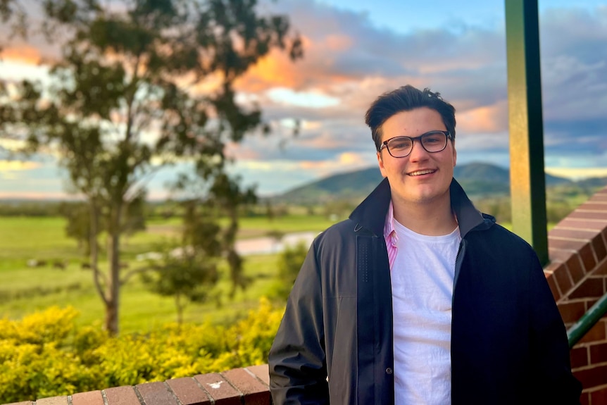 a young man in a white shirt and black jacket stands in front of a sunset