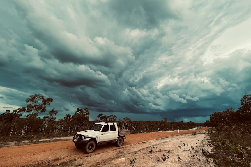 A white ute is parked on a dirt road with trees and dark clouds behind it