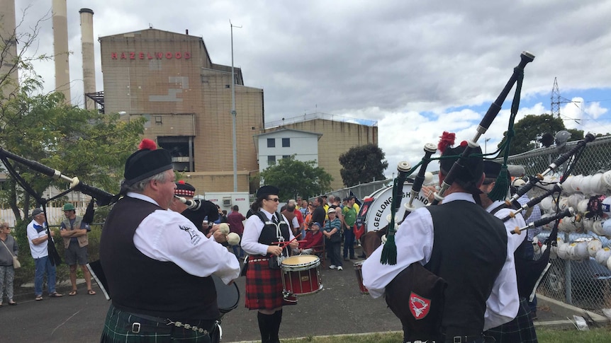A group of people wearing kilts and traditional Scottish dress play bagpipes for a crowd outside the Hazelwood power station.