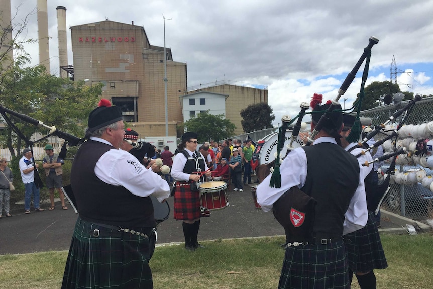 A group of people wearing kilts and traditional Scottish dress play bagpipes for a crowd outside the Hazelwood power station.