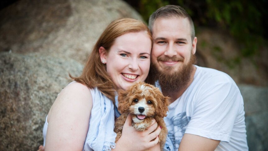 A woman smiles big while holding a brown puppy, sitting next to a man with a beard who is smiling