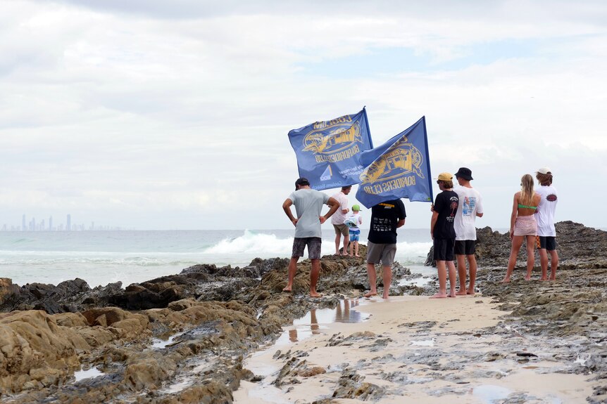 People with flags on a rock at the beach watching surfers. The Gold Coast skyline is in the background.