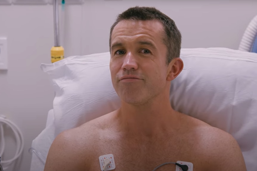 A shirtless man smiles proudly at the camera from a hospital bed.