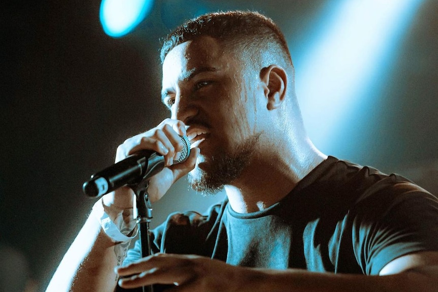 A young Pacific Islander man with a faded haircut raps into a microphone as sweat drips down his face under blue stage lighting