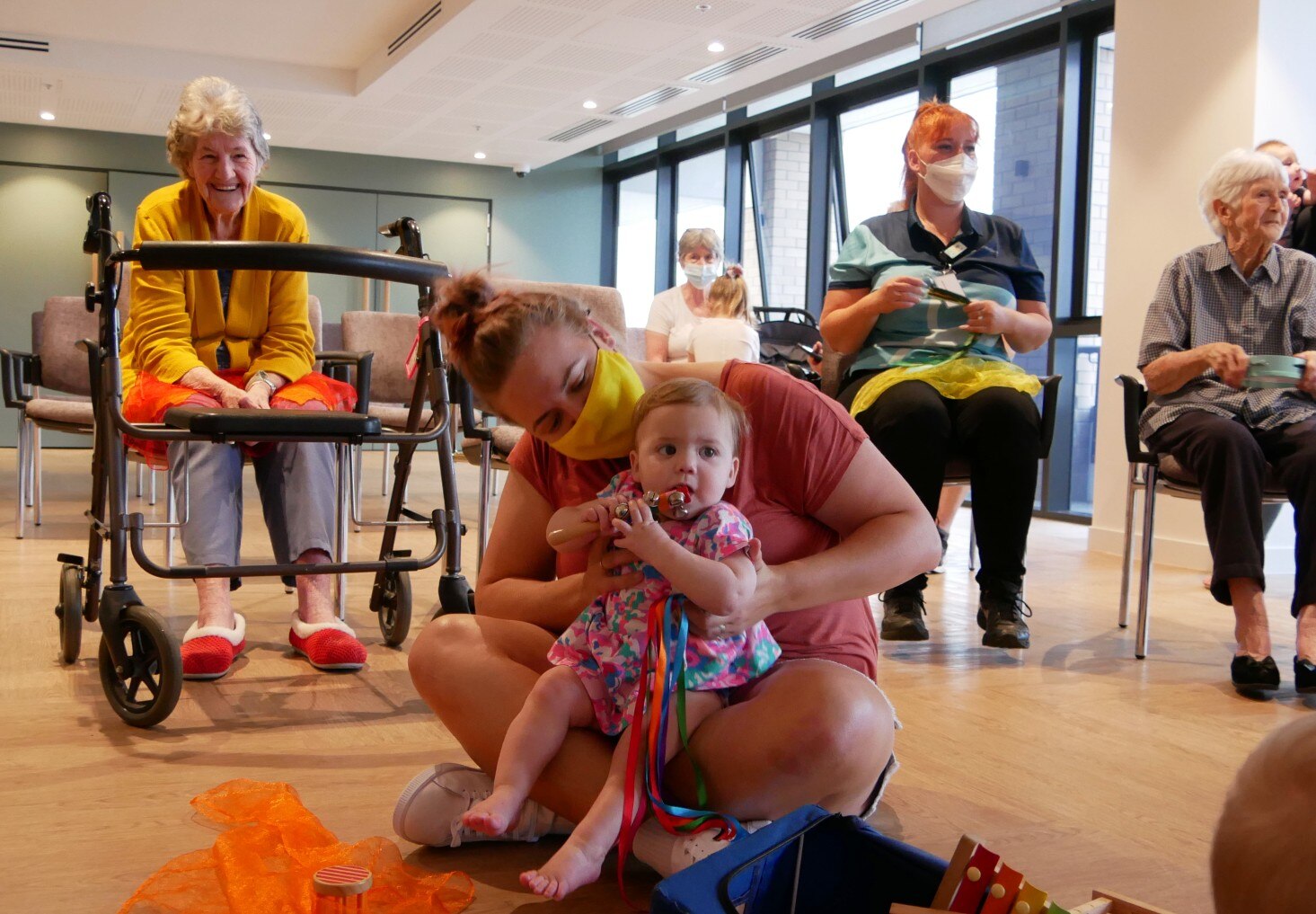 A woman with a baby on her lap, in the background a walking frame and an elderly woman smiling
