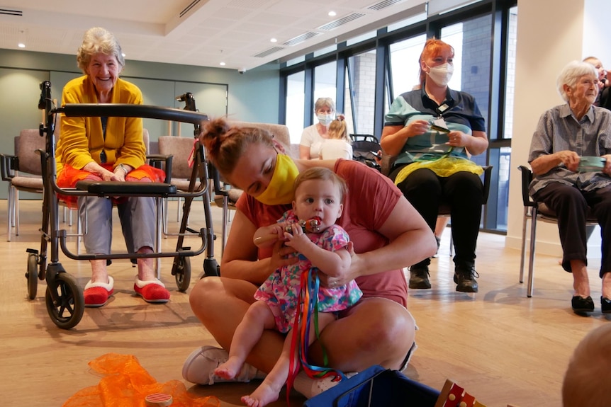 A woman with a baby on her lap, in the background a walking frame and an elderly woman smiling