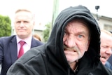 a man in a dark hoodie looking angry as he is flanked by reporters