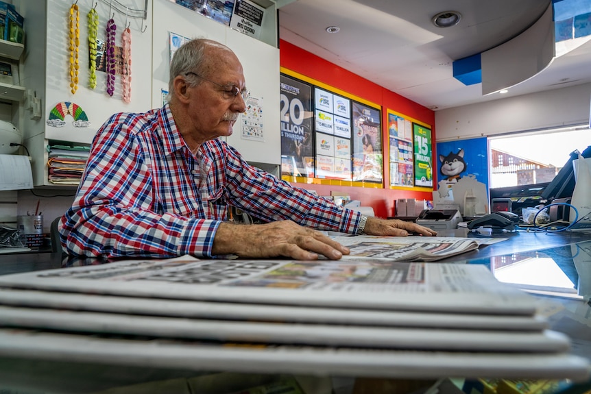 A low shot of a man reading a newspaper at a counter with a stack of newspapers in the foreground.