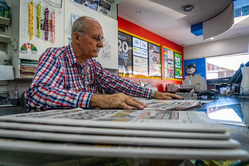 Un tiro bajo de un hombre leyendo un periódico en un mostrador con una pila de periódicos en primer plano.