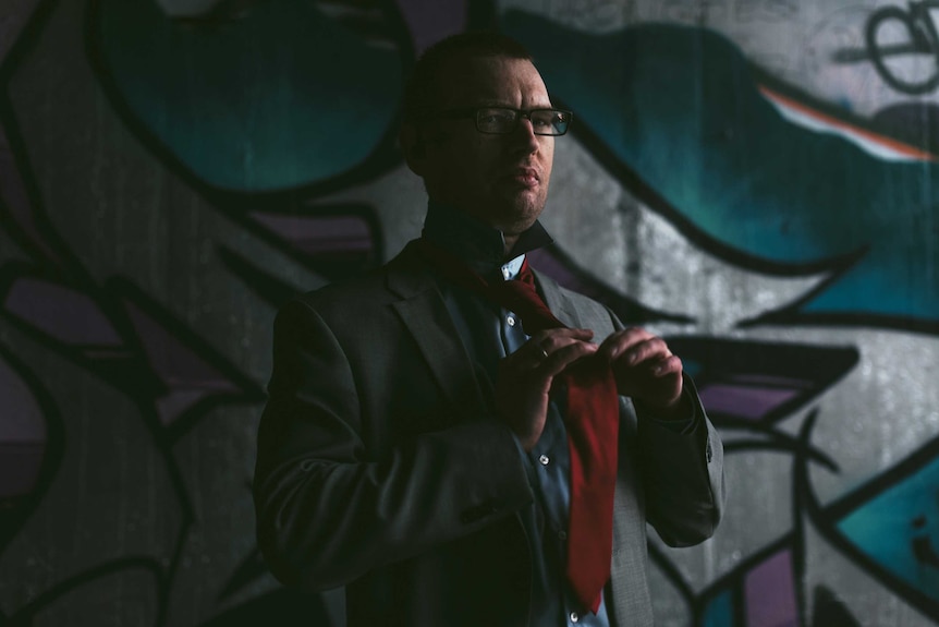 Christopher Anderson tying a tie, standing in the dark in front of a wall with graffiti