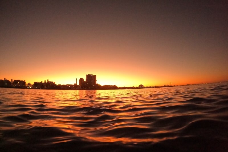 The sun rising over the dark water with buildings in the background.