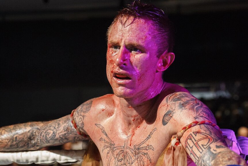 A man with tattoos and no shirt leans against the ropes of a boxing ring