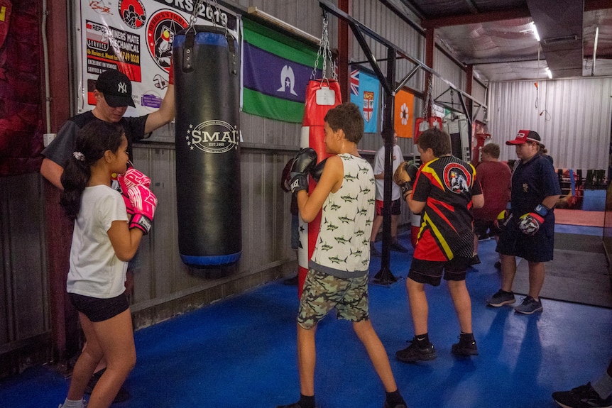 A group of girls and boys training with punching bags in a colourful gym. They are overseen by an older instructor
