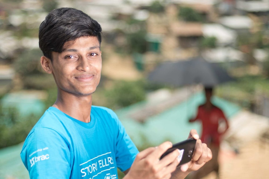 A boy in a blue t-shirt holds his mobile phone and smiles at the camera.