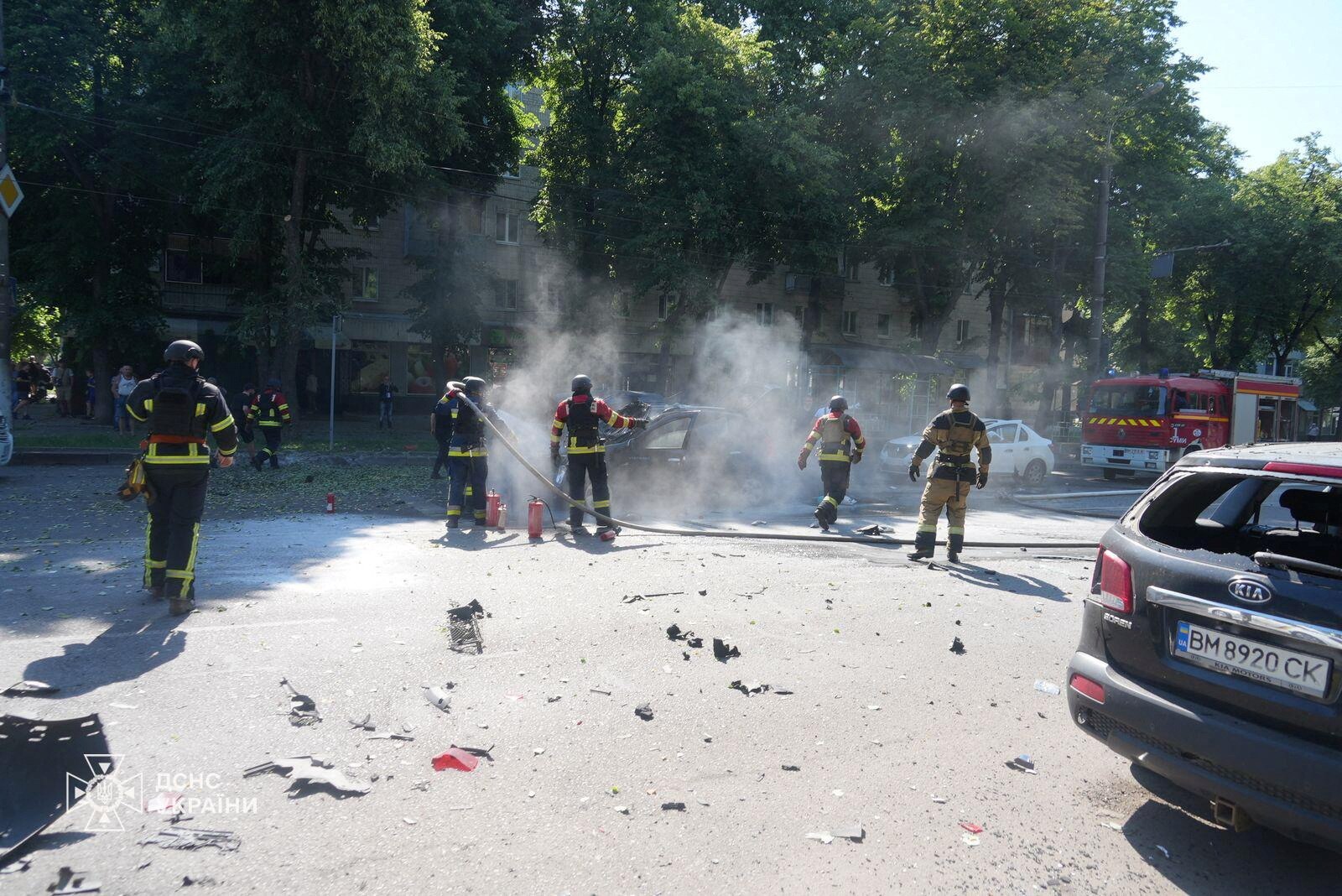 Firefighters stand on a street with cars and debris. 