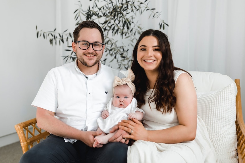 A man and woman smiling and holding a baby while sitting no a couch.