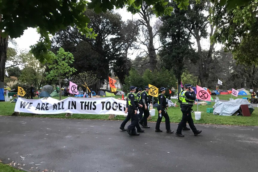 A group of police in high-vis vests walk past a makeshift campground and a sign saying 'we are all in this together'.