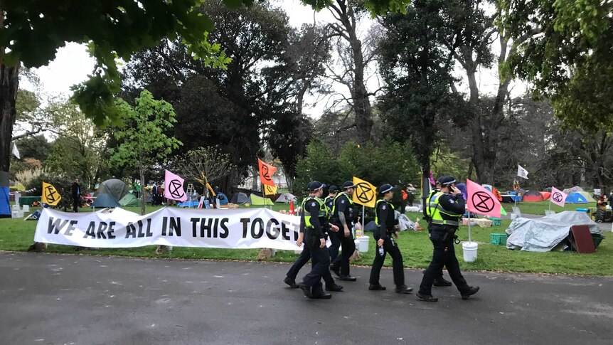 A group of police in high-vis vests walk past a makeshift campground and a sign saying 'we are all in this together'.