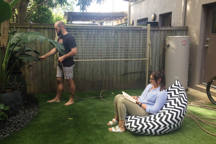 A couple in their small backyard, lady relaxes while man hoses plants.