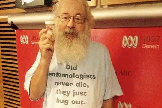 An older, bespectacled man with a long white beard holding up a small jar containing an insect.