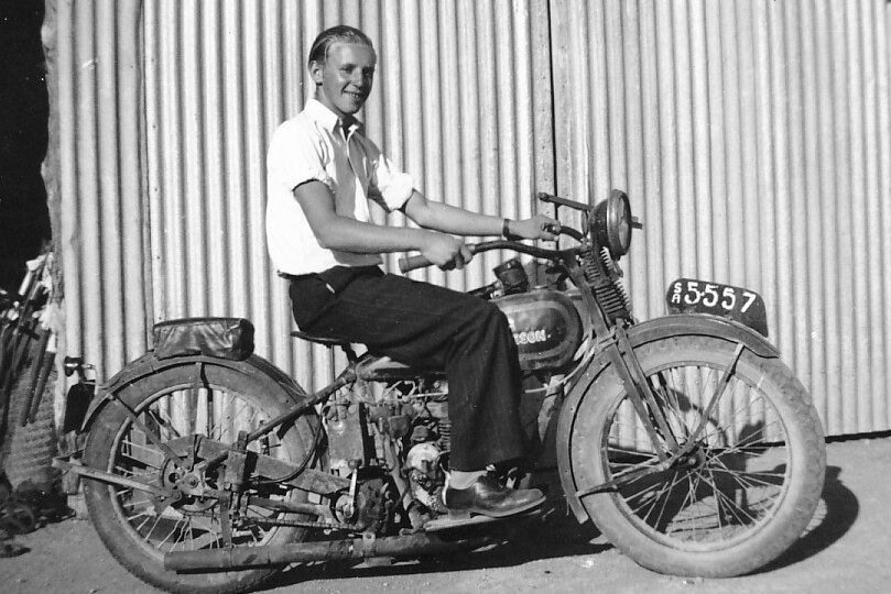 Well dressed man in white collared shirt and black pants on antique motorbike in front of corrigated iron shed
