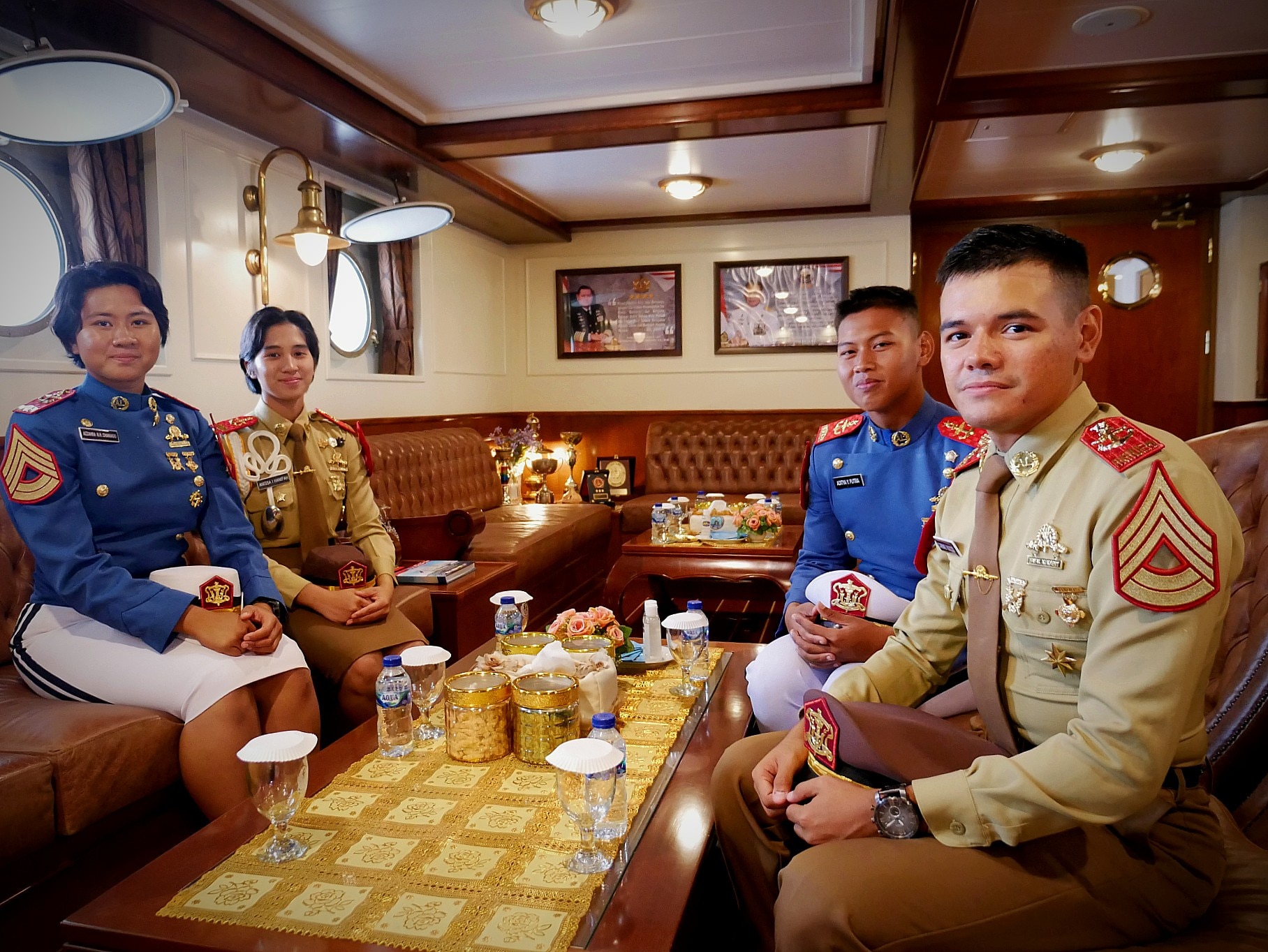 Four naval cadets in blue and green dress uniforms sit down in a ship's ballroom with wooden furniture and gold details.