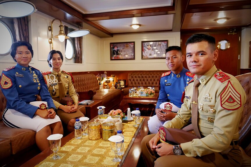 Four naval cadets in blue and green dress uniforms sit down in a ship's ballroom with wooden furniture and gold details.