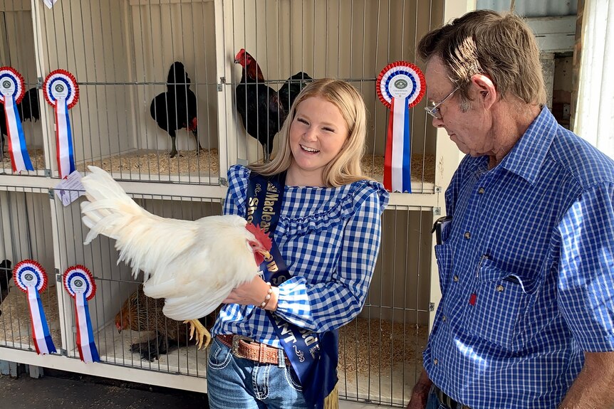 A teenage girl in a blue and white check shirt holds a white rooster next to a man in a blue check shirt.