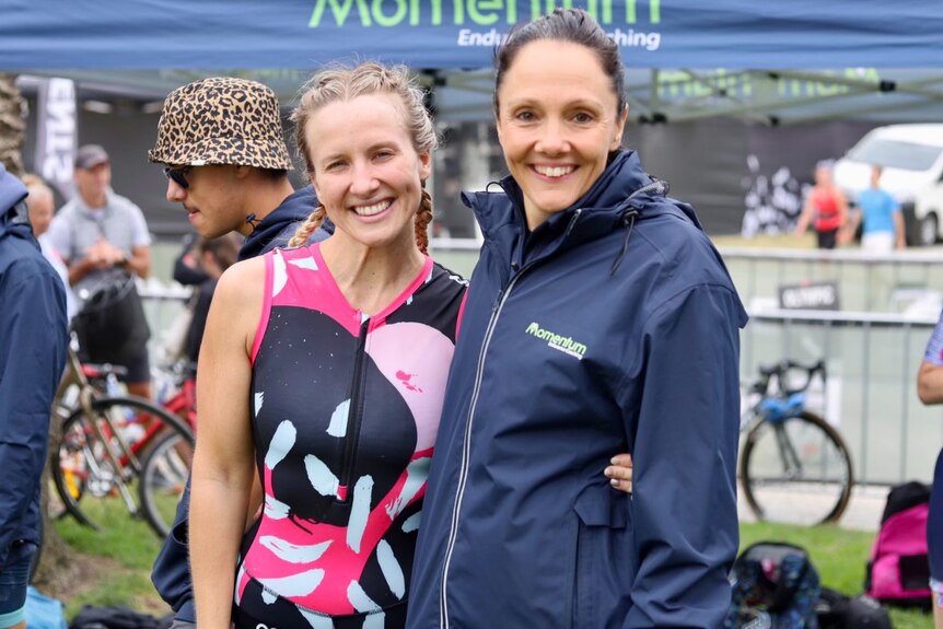 Samantha Janssen wearing a navy sports parka with her arm around a triathlon athlete, both smiling and looking at the camera