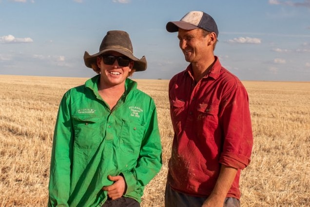 Two farmers standing in paddock, both smiling. One wears green shirt, large hat, other red shirt, cap.