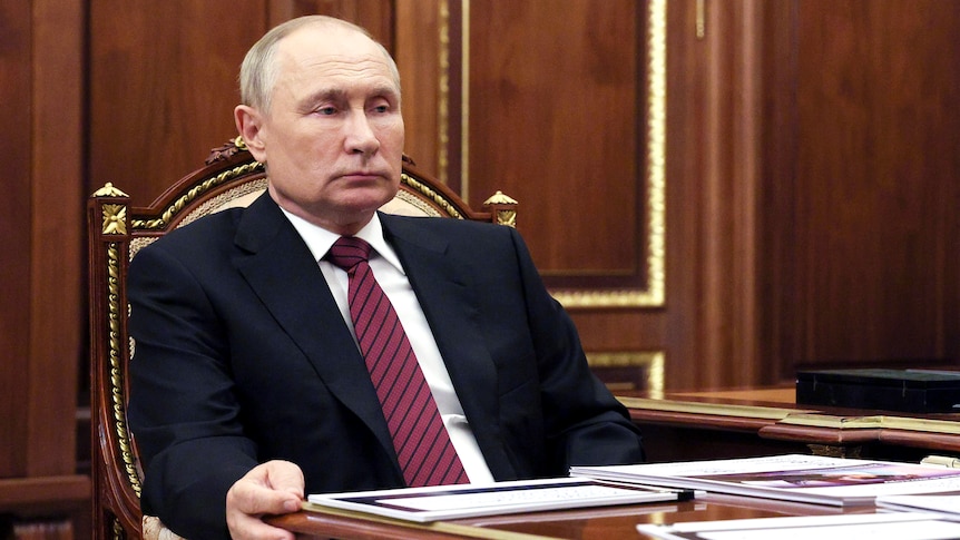 Vladimir Putin sits at a desk with a very ominous expression on his face.