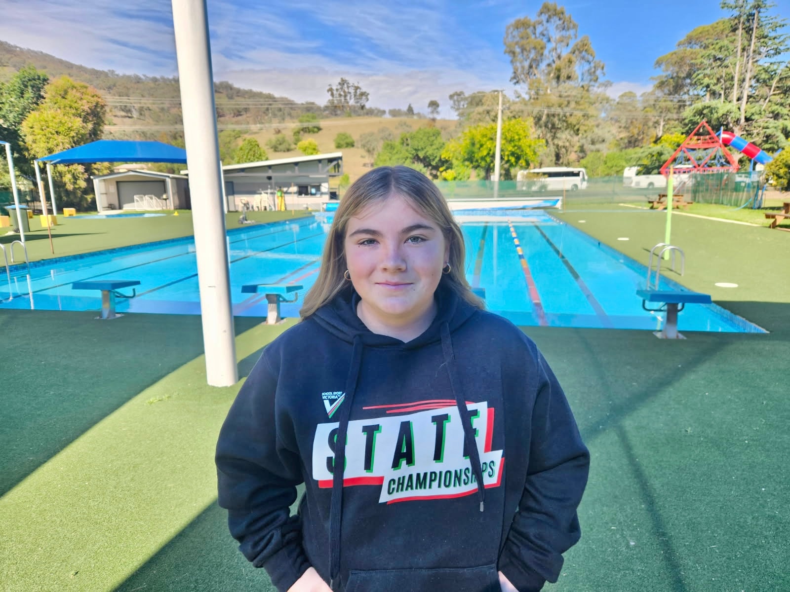 A young girl wearing a black hooded jumper standing on a green surface in front of an outdoor pool