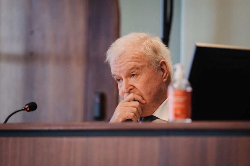 A man with white hair sits behind a desk with his hand over his mouth.