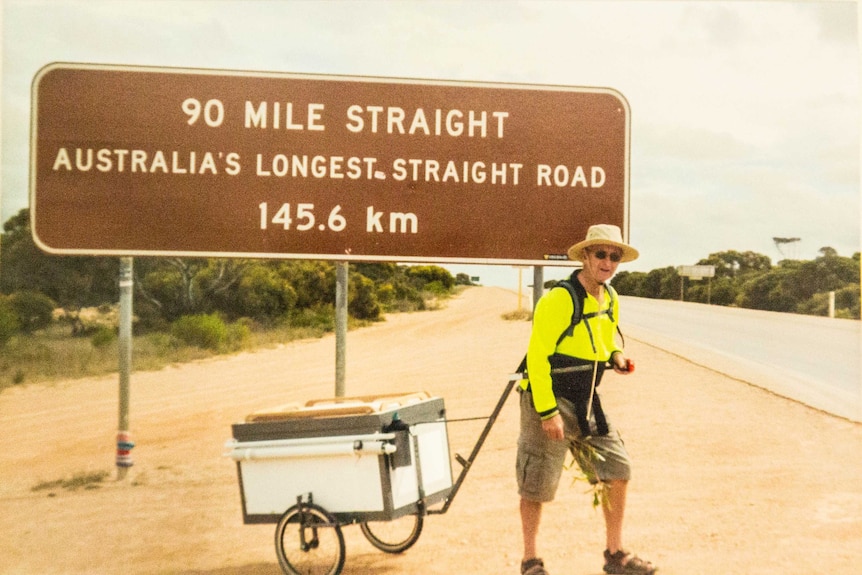 An archival photo of Mr Burgess standing on the Nullarbor Plains with a large sign behind him.