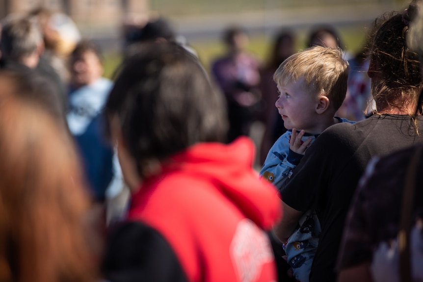 A young boy being held in an adult's arms looks out over a crowd of people.