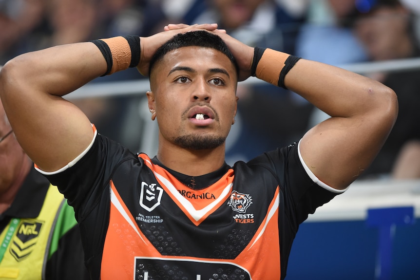 A Polynesian Rugby player in orange and black kits look on with anguish from sideline of packed stadium with hands on head