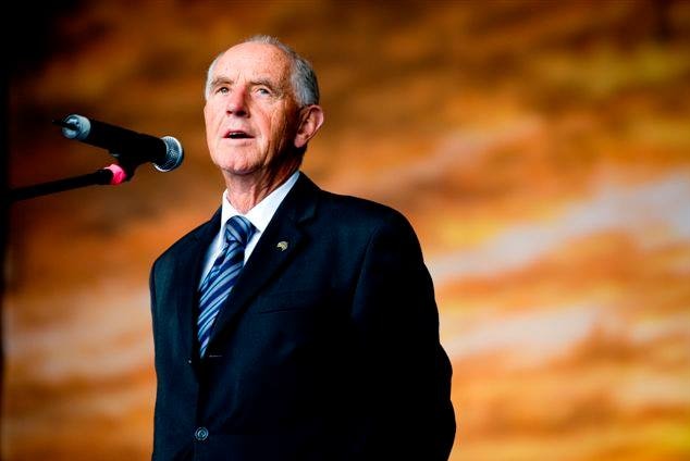 An elderly man wearing a suit and tie speaks into a microphone against a blurry orange backdrop.